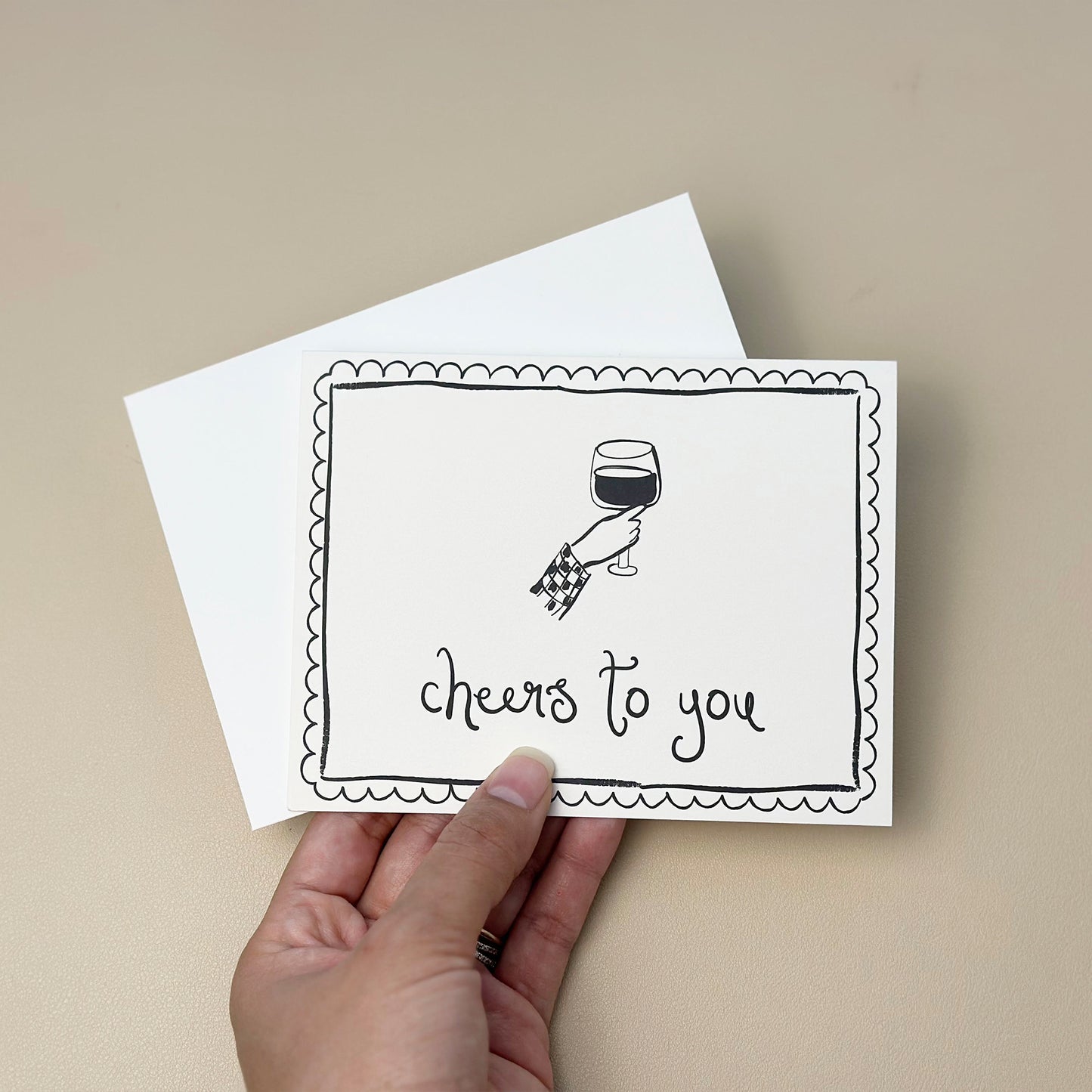 A minimalistic, hand-drawn greeting card with the text "cheers to you," featuring an illustrated hand holding a wine glass. The card has a scalloped border design and comes with a matching white envelope. Perfect for celebrations, congratulations, and special occasions.
