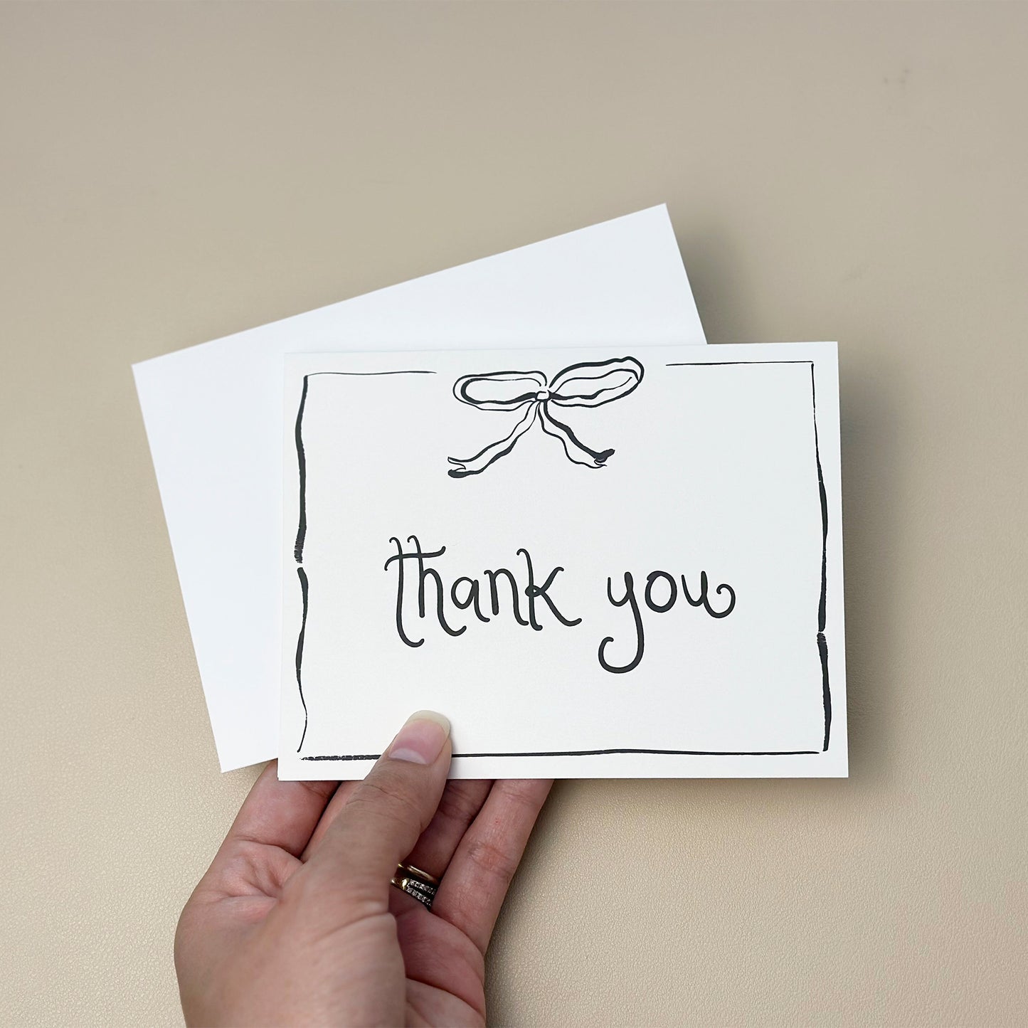 A minimalist thank you card featuring a hand-drawn bow illustration at the top and the phrase "thank you" written in a whimsical font. The card includes a delicate border design and comes with a white envelope, perfect for expressing gratitude for any occasion.