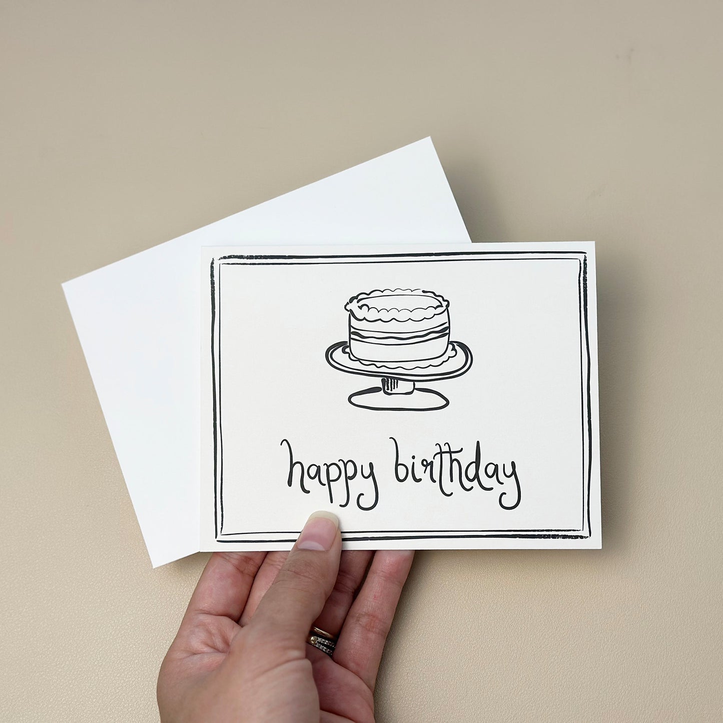 A hand-drawn birthday card featuring an illustration of a layered cake on a cake stand, paired with the phrase "happy birthday." The card has a clean border design and comes with a white envelope, perfect for birthday celebrations and well wishes.