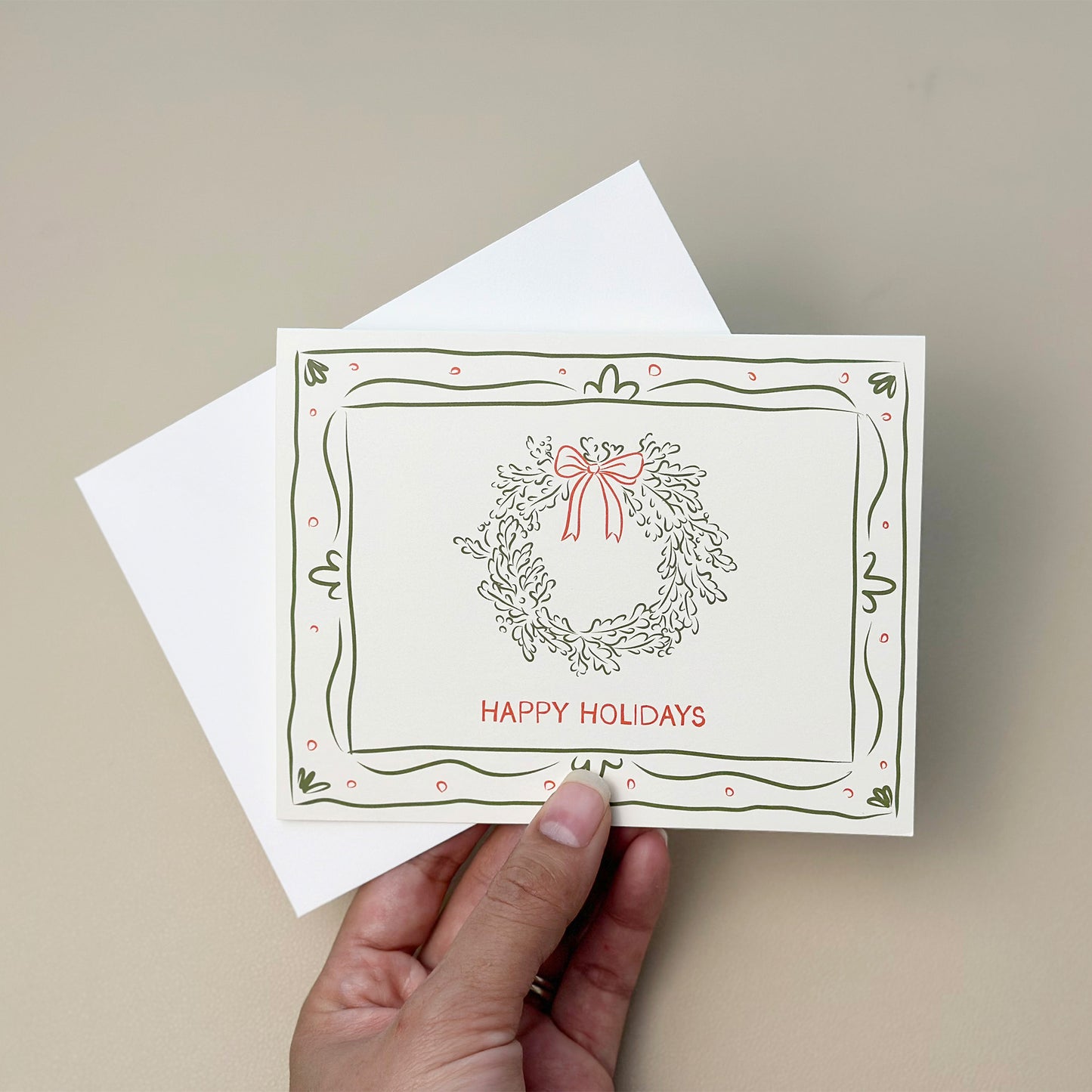 A festive "Happy Holidays" card featuring a hand-drawn wreath with a red bow and green border design. This charming holiday greeting card includes a matching white envelope, perfect for spreading joy and warm wishes during the holiday season.