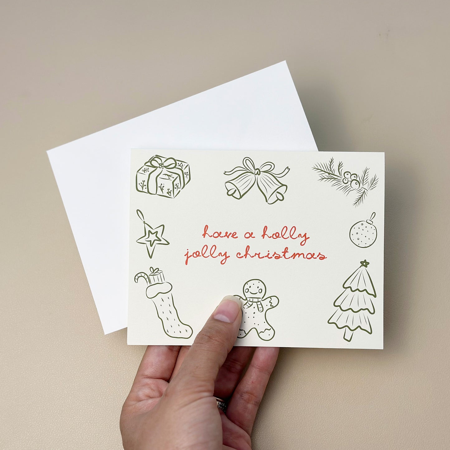 A festive "Holly Jolly Christmas" card with playful hand-drawn illustrations of holiday elements like a gingerbread man, bells, stockings, and pine branches. The card is designed with red text and comes with a matching white envelope, ideal for spreading holiday cheer.