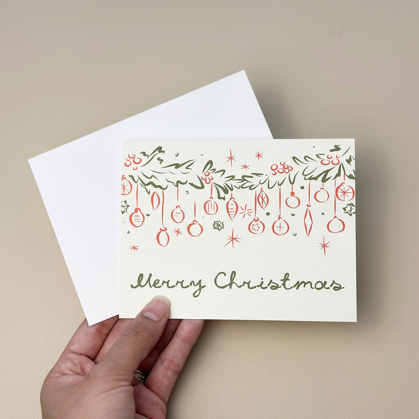 A festive "Merry Christmas" card featuring hand-drawn garland, ornaments, and holly berries in green and red line art. The card includes elegant typography and comes with a matching white envelope, perfect for sharing holiday cheer.