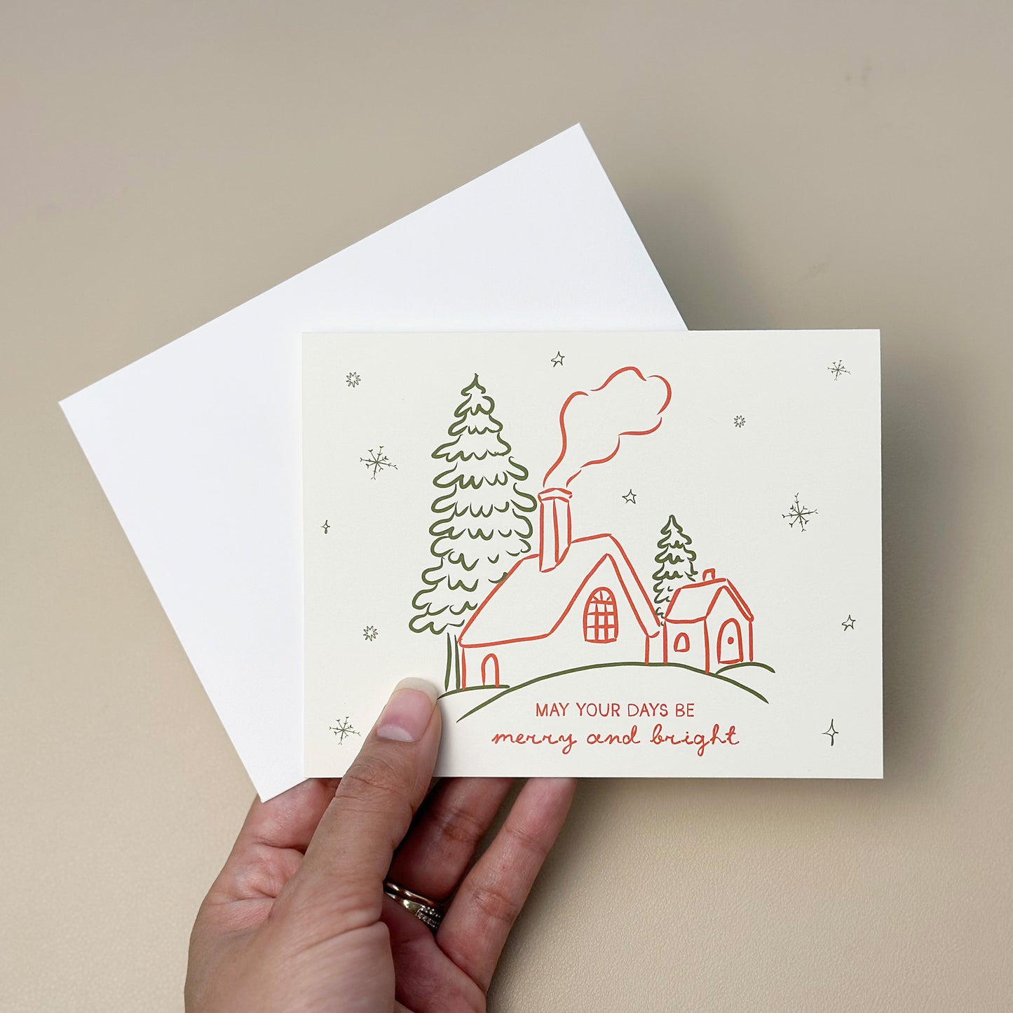 A charming "Merry and Bright" greeting card with whimsical hand-drawn illustrations of a snow-covered cottage and trees. Designed with festive red and green line art, this card comes with a matching white envelope, ideal for sending warm holiday wishes.