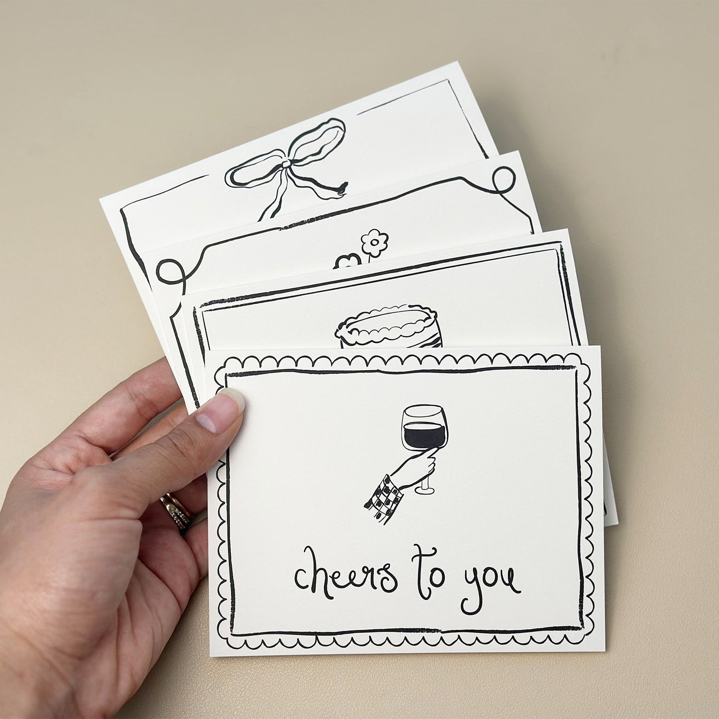 A collection of four hand-drawn greeting cards from "The Essentials Set," featuring designs for "thank you," "cheers to you," "happy birthday," and "thinking of you." Each card includes whimsical line art with minimalistic illustrations and a clean border design, paired with matching white envelopes.