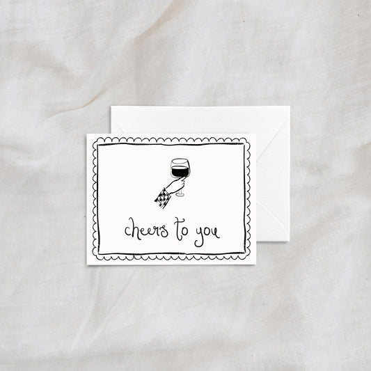A stylish and modern greeting card with the phrase "cheers to you," showcasing a hand-drawn illustration of a hand holding a wine glass. The card includes an elegant scalloped border and is paired with a white envelope, ideal for weddings, anniversaries, or celebratory messages.