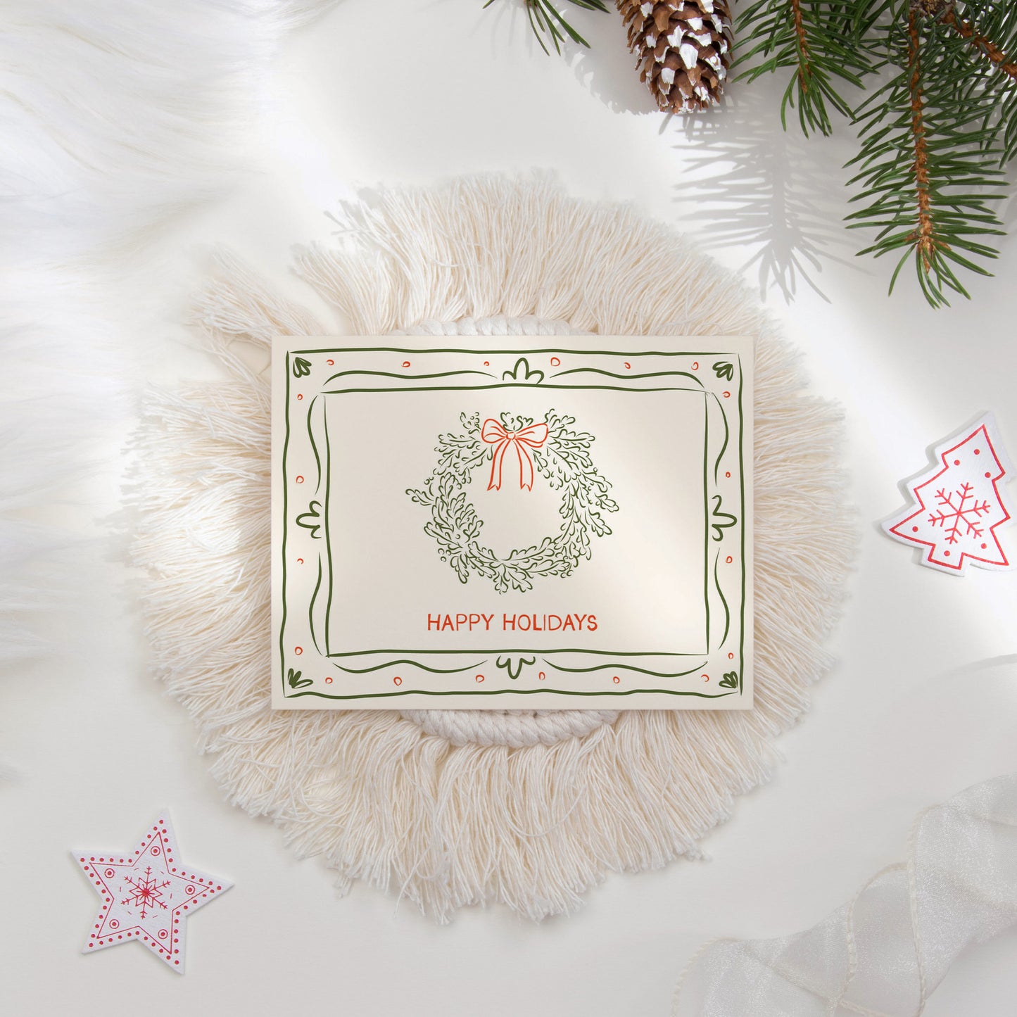 A beautifully styled flat lay of a "Happy Holidays" card, adorned with a hand-drawn wreath illustration and red bow. The card features green decorative borders and is surrounded by festive elements like pine branches, ornaments, and a cozy textured mat, ideal for holiday gifting and seasonal greetings.