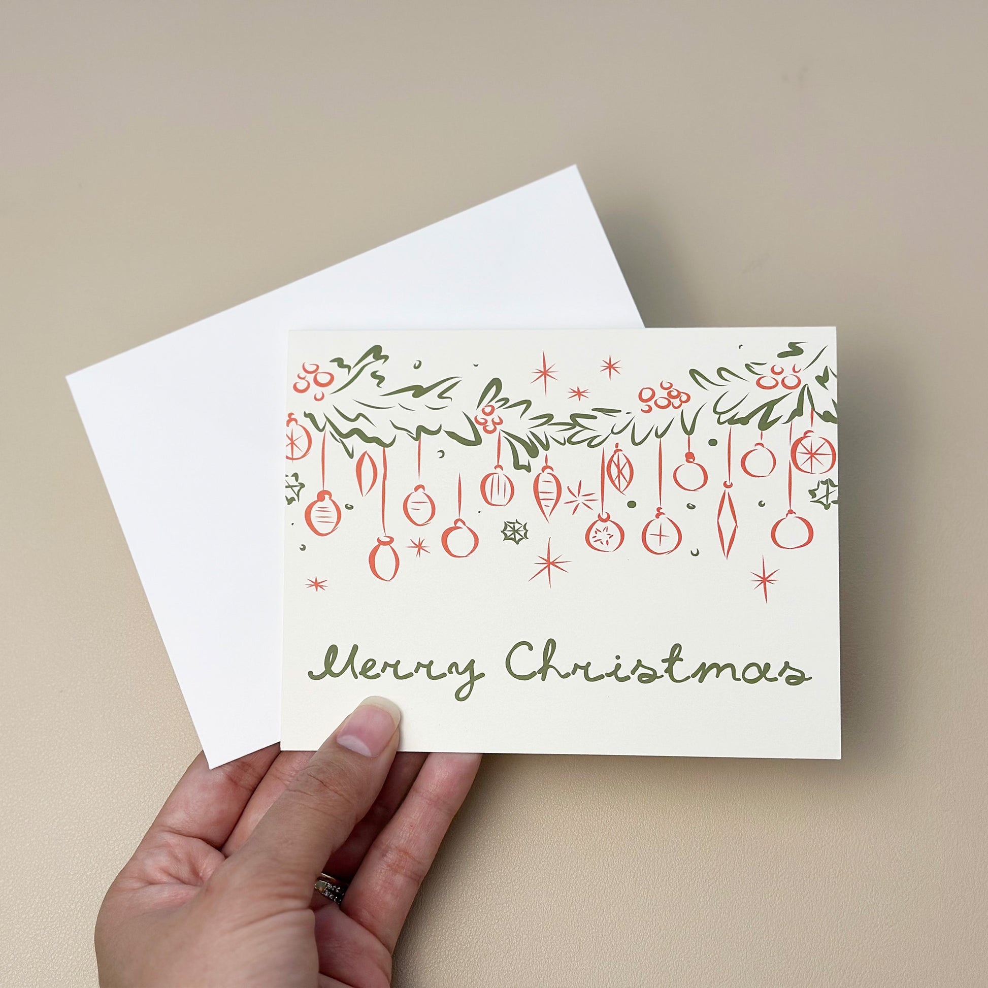 A festive "Merry Christmas" card featuring hand-drawn garland, ornaments, and holly berries in green and red line art. The card includes elegant typography and comes with a matching white envelope, perfect for sharing holiday cheer.