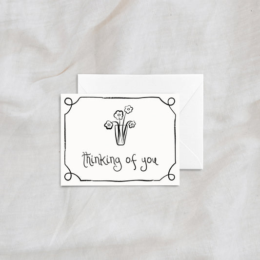 A charming "thinking of you" greeting card with a simple floral vase illustration and decorative border. Designed with clean line art and paired with a matching white envelope, this card is ideal for thoughtful and comforting notes.