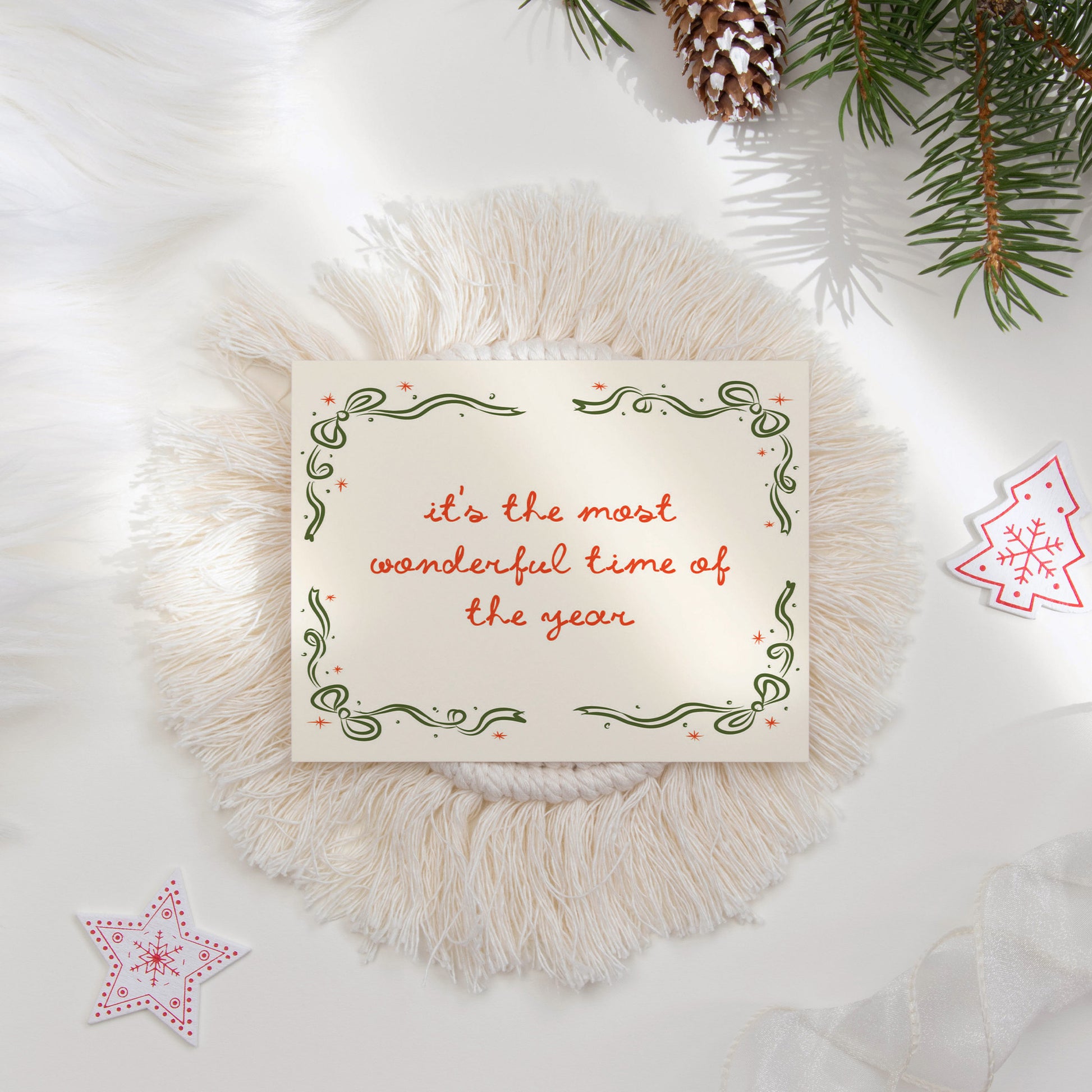 A festive "It's the Most Wonderful Time of the Year" holiday card featuring a hand-drawn green border with decorative swirls and red stars. The flat lay is styled with cozy holiday elements like pine branches and textured decor, perfect for seasonal greetings.