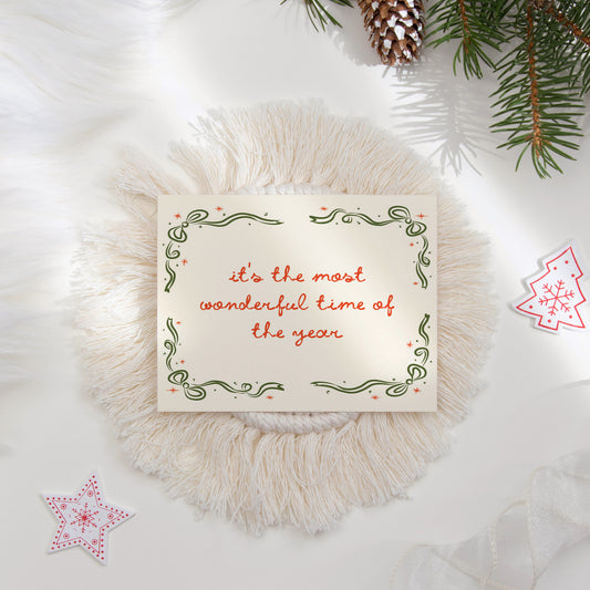 A festive "It's the Most Wonderful Time of the Year" holiday card featuring a hand-drawn green border with decorative swirls and red stars. The flat lay is styled with cozy holiday elements like pine branches and textured decor, perfect for seasonal greetings.